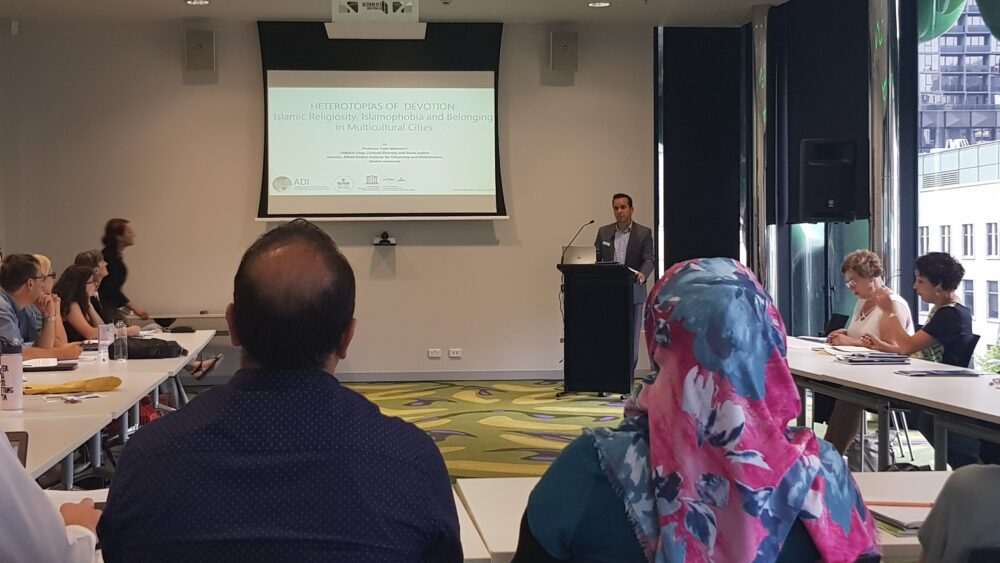 Fethi Mansouri standing at a podium at the front of a large room, with a presentation projected onto the wall, addressing people sitting around a U shaped table.