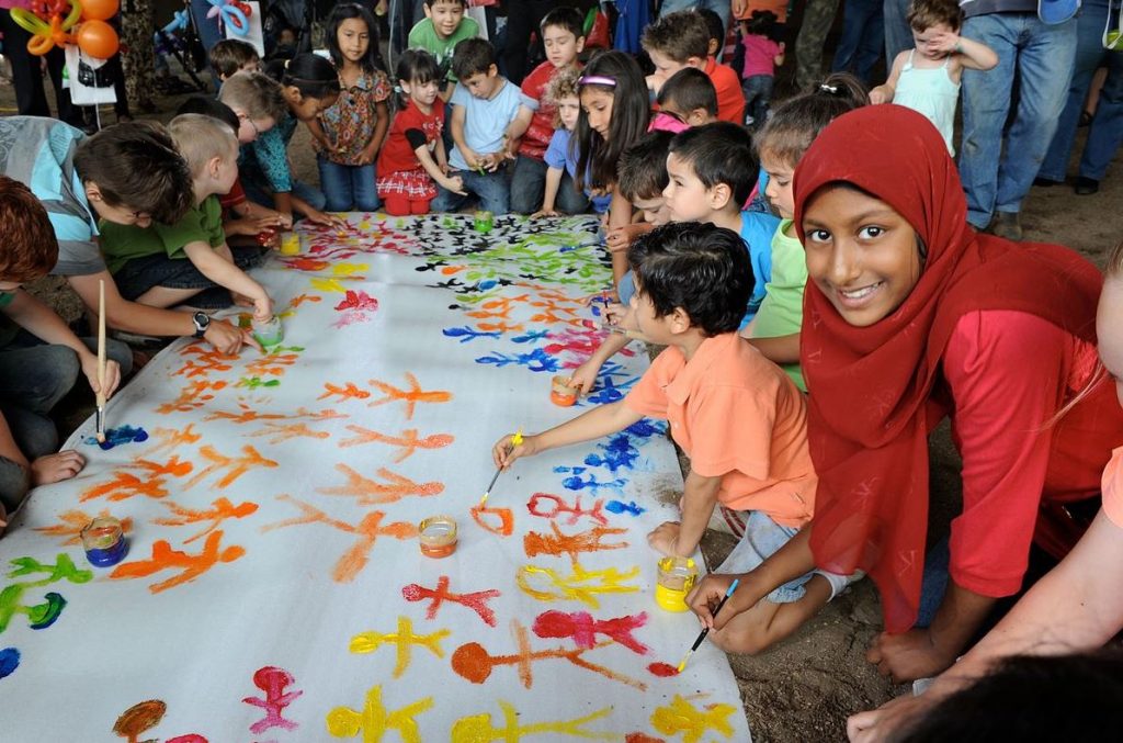 A large group of happy children from diverse ethnic backgrounds crouching down together, painting a long mural of many different coloured stick figures on paper. One girl on the right with big brown eyes and brown skin, wearing a red hijab, is looking at the camera and smiling.