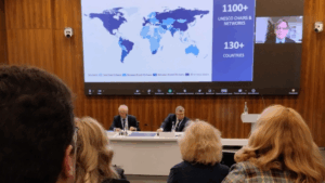 A brightly lit conference room where the 5th Congress of Russion UNESCO Chairs is being held, where a large map of the world is being displayed on a slide deck while a man speaks to the audience via video. The audience appears to be full.