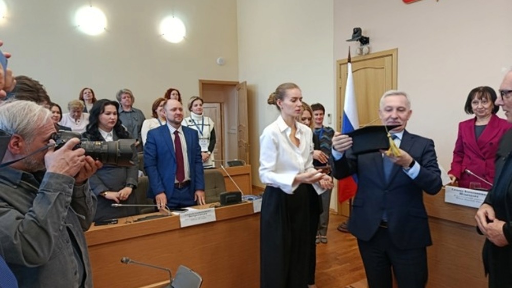 An image of a well lit and crowded lecture hall, where an international conference is being help in St Petersburg. In the centre, a man holds an academic cap which he appears to be presenting to someone else. Everyone is standing to watch and a photographer is present.